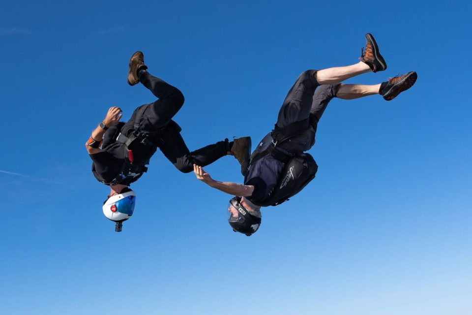 Two skydivers angle flying with one placing his food on the chest of the other simulating sky surfing