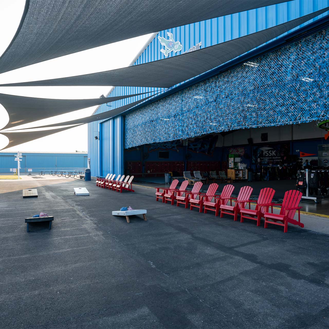 Cornhole boards and shaded outdoor seating area with red adirondack chairs at Start Skydiving
