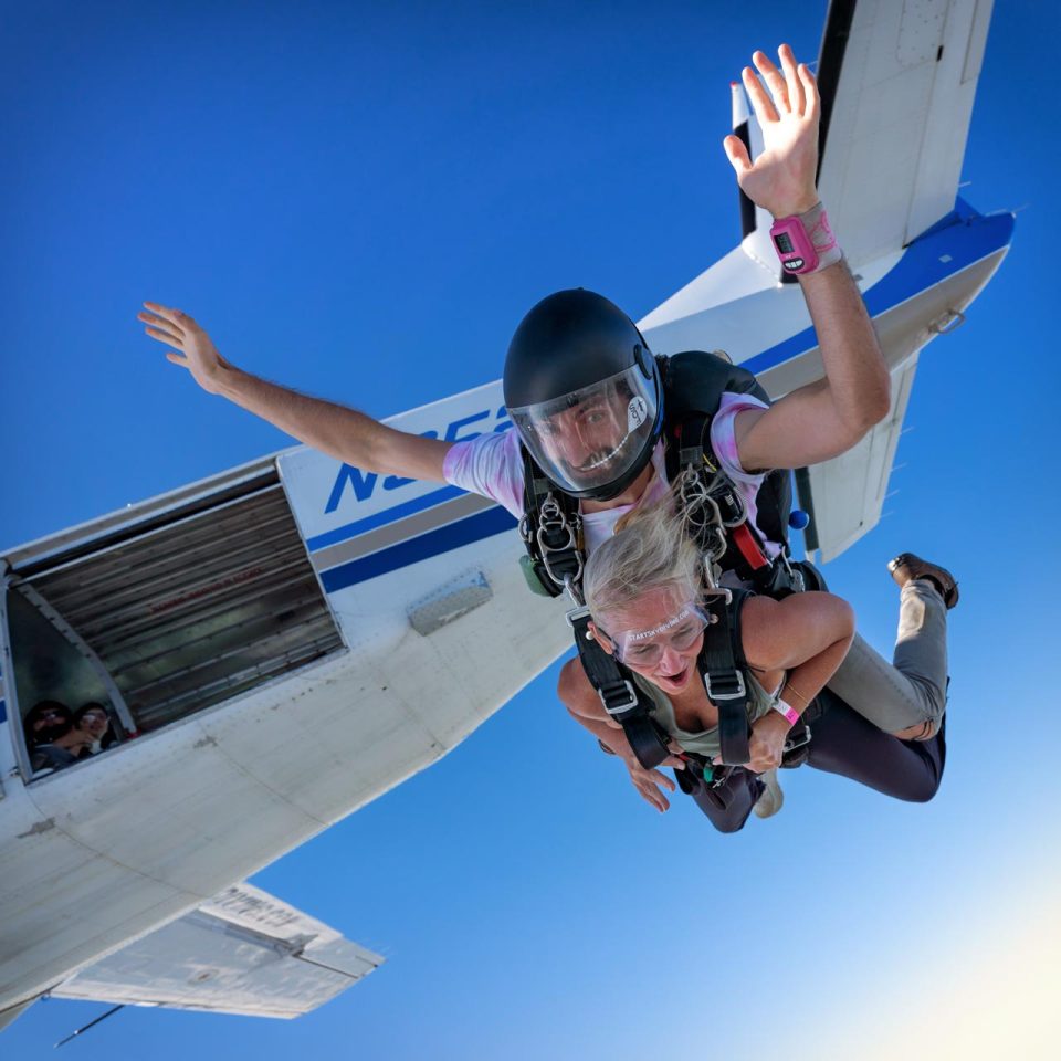 Male tandem instructor with a black full face helmet smiles with his hands up as he and his tandem student exit the aircraft.