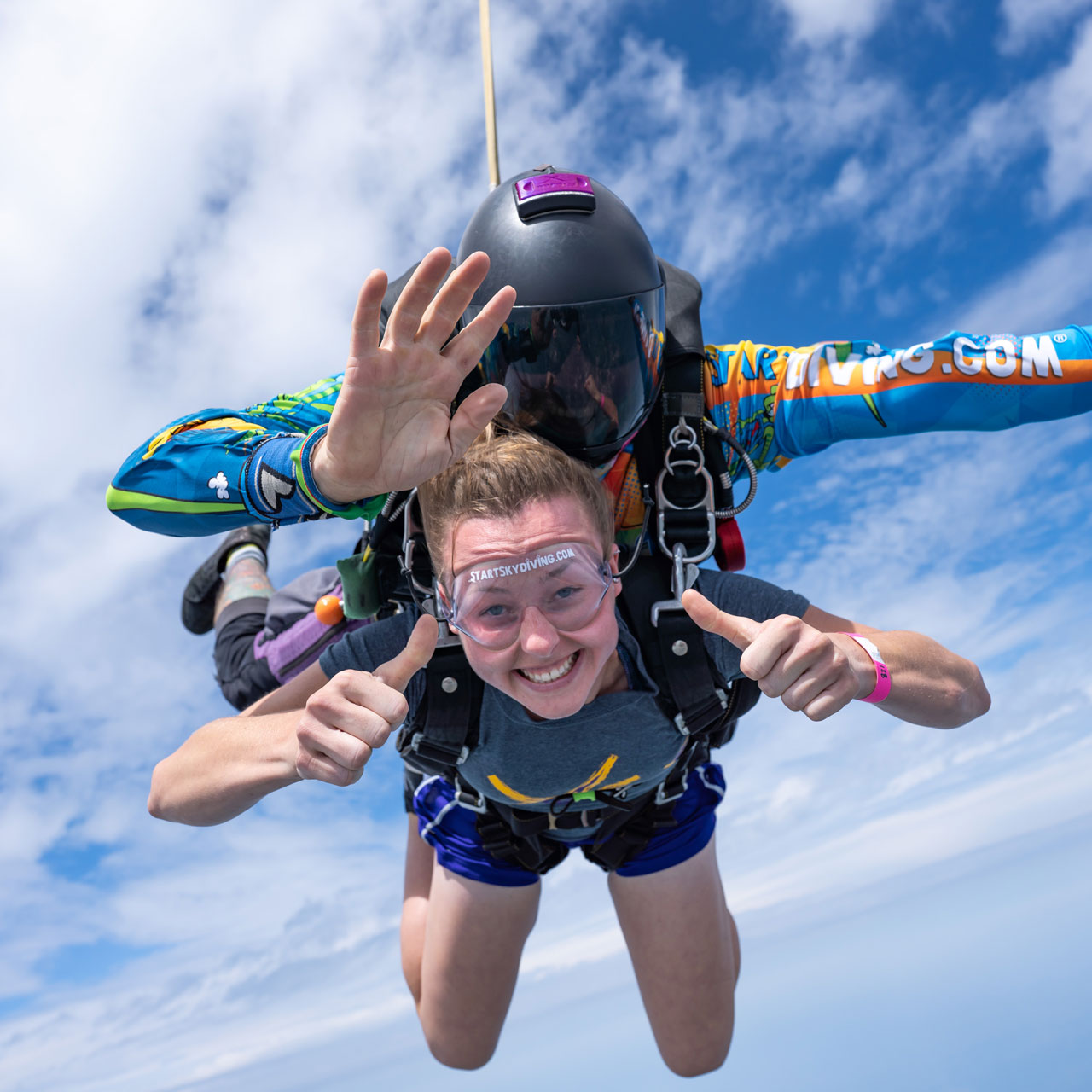 Female tandem student gives thumbs up wearing shorts and short sleeved during freefall with her instructor.